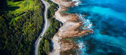 Aerial view of a coastal road curving between a lush green landscape and a rocky shoreline with clear blue ocean waters.