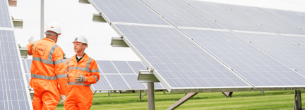 Two workers in orange safety uniforms and helmets inspecting large solar panels at a solar farm on a sunny day.